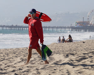 Lifeguard on the beach facing the camera wearing Nöz reef safe sunscreen on his nose carrying swim fins and a rescue board